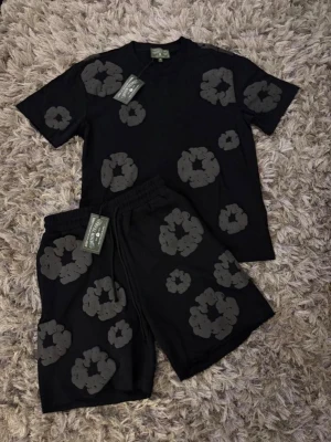 Denim Tears shorts med blommigt tryck - Shorts från Denim Tears med elastisk midja och snörning. Finns i svart med grått eller vitt blommigt tryck samt i ljusrosa med vitt tryck. Avslappnad passform och tillverkade i mjuk bomull, perfekta för sommaren.