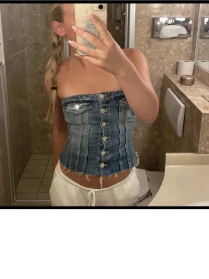 Blå denim tubtopp med knappar - Säljer en snygg tubtopp i blå denim med silverfärgade knappar framtill och två fejkade bröstfickor. Toppen är croppad med rå kant nedtill för en cool vibe. Perfekt för dig som vill sticka ut med en unik jeanslook.