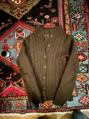 Brun kabelstickad kofta från Tommy Hillfiger - Mysig brun kabelstickad kofta från Hilfiger med grov ribbad krage och knappar framtill. Snygga flätade detaljer längs hela tröjan och långa ärmar. Perfekt för dig som gillar klassisk stil med lite extra edge. Är i storlek M men sitter verkligen som en kofta i XS