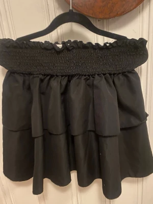 Svart volangkjol med smock - Supersöt svart kortkjol med bred smock i midjan och dubbla volanger. Kjolen har en luftig känsla och passar perfekt till en trendig look. Den elastiska midjan gör den bekväm och lätt att bära. Perfekt att matcha med en crop top eller stickad tröja.