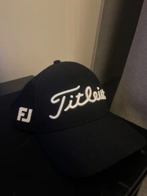 Svart Titleist keps FJ - Snygg svart keps från Titleist med vit broderad logga framtill och detaljer som FJ och Pro V1 på sidorna. Insidan har svarta band med Titleist-tryck och texten Tour Performance. Justerbar baktill med röd söm. Perfekt för golfbanan eller chill vardagsstil.