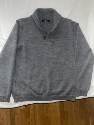 Grå stickad tröja  - Säljer en grå stickad tröja från Zara Man i storlek L. Tröjan har en bred krage med en knappdetalj och ribbade muddar vid ärmslut och nederkant. Perfekt för kyliga dagar och enkel att matcha med jeans eller chinos.