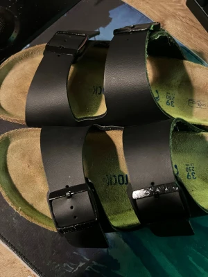 Svarta Birkenstock sandaler strl 39 - Birkenstock sandaler med två breda svarta remmar i skinn och justerbara spännen. Fotbädden är ergonomiskt utformad i beige mocka och sulan har ett unikt mönster för extra grepp. Perfekt för chill dagar och sommarhäng.