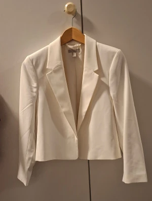 Vit kavaj från H&M, storlek XS - Snygg vit kavaj från H&M i storlek XS. Klassisk design med krage och lång ärm, croppad passform och stilrena linjer. Perfekt för dig som gillar minimalistisk och clean stil. Materialet känns mjukt och har en lätt glansig finish.