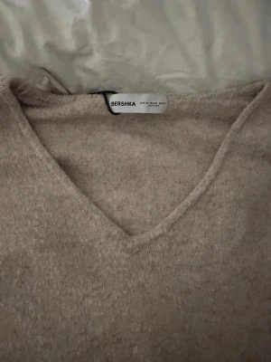 Beige stickad v-ringad tröja Bershka - Mysig beige stickad tröja från Bershka med v-ringad hals och ribbade muddar vid ärmslut och nederkant. Perfekt för lager-på-lager och chill dagar. Långärmad och i en loose passform som ger en avslappnad vibe.