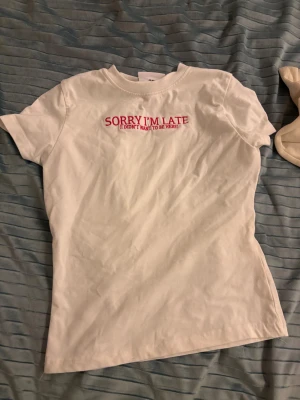 Vit t-shirt med röd text - Vit t-shirt med korta ärmar och rund halsringning. På bröstet står det 'SORRY I'M LATE (I DIDN'T WANT TO BE HERE)' broderat i rött. Skön och enkel modell i mjuk bomull, perfekt för en chill och avslappnad stil.