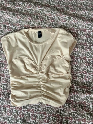 Beige croppad topp med scrunch från cider - Säljer en beige croppad topp från cider  med snygg rynkdetalj framtill och bak. Toppen har korta ärmar och en söt sweetheart-liknande söm över bysten. Mjuk och stretchig i bomullsmix, perfekt till fest 