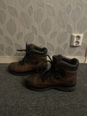 Bruna skinnkängor med snörning - Stabila kängor i brunt skinn med svarta detaljer och kraftig sula. De har klassisk snörning med metallöglor och vadderad kant runt ankeln för extra komfort. Perfekta för dig som gillar outdoor och vill ha en robust look.