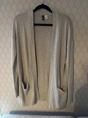 Ljusgrå stickad kofta från Divided - Mysig beige stickad kofta från Divided med öppen front och två stora fickor framtill. Perfekt att slänga på sig över en t-shirt eller topp. Långärmad och med ribbade muddar för extra komfort.