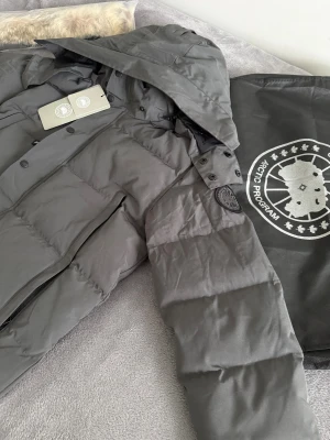 Canada Goose jacka  - Snygg grå dunjacka från Canada Goose med svart logotyp på ärmen och avtagbar huva. Jackan har raka linjer, stora fickor med dragkedja och knappar framtill. Perfekt för kalla dagar och har en klassisk, stilren look.