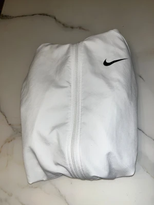 Vit Nike vindjacka med huva - Snygg vit Nike vindjacka med huva och hel dragkedja framtill. Jackan har Dri-FIT-material som andas och är perfekt för aktiva dagar. Svart Nike-logga på bröstet ger en clean och sportig look. Fickor på sidorna och stilren passform.