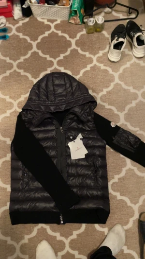 Svart Moncler pufferjacka med huva - Svart pufferjacka från Moncler med huva och stickade svarta ärmar. Jackan har dragkedja framtill och på ärmen, samt Moncler-logga på vänster ärm. Materialmix av polyester och stickat, quiltad kropp och praktisk ficka på ärmen. Snygg och modern look. Fraktas inom 24h 