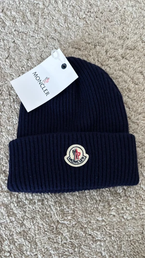 Mörkblå mössa - Snygg mörkblå mössa med ribbstickad design och klassisk logga framtill. Mössan är tillverkad i mjukt stickat material och har en bred uppvikt kant. Perfekt för att hålla värmen och ge din outfit en lyxig touch.