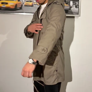 Beige parkajacka med huva och foder - Säljer en beige parkajacka med huva och mysigt foder på insidan. Jackan har knappar, dragkedja och snörning i midjan för snygg passform. Flera fickor framtill och detaljer vid ärmslut. Perfekt för kyliga dagar.