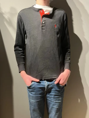 Svart långärmad tröja Polo Ralph Lauren - Svart långärmad tröja från Polo Ralph Lauren med röd broderad logga på bröstet. Tröjan har en rund hals med knappslå och röda detaljer vid knapparna. Tillverkad i mjuk bomull, perfekt för dig som gillar stilrena och klassiska plagg.