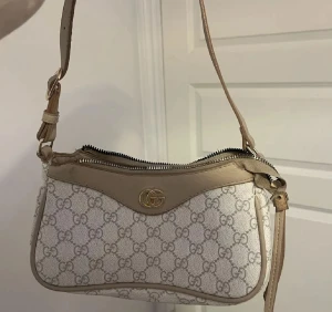 Beige axelväska  - Säljer den så billig då kedjan är trasig ( man får fixa det själv , det går att fixa. ) Snygg beige axelväska med klassiskt GG-mönster och guldfärgad logga framtill. Väskan har dragkedja upptill och justerbar axelrem i skinn. Perfekt storlek för det viktigaste och en stilren look.