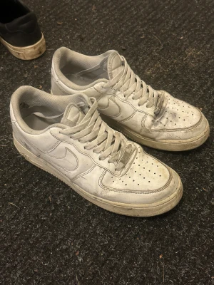 Nike Air Force 1 vita sneakers - Klassiska Nike Air Force 1 sneakers i vitt skinn med perforerad tå och rundad form. Skorna har snörning och platt sula, samt tydlig Nike-logga på sidan och hälen. Perfekt för dig som gillar streetstyle och tidlös design.