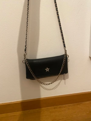 Svart axelväska - Svart väska från Gina tricot,den har en jätte gullig liten stjärna i mitten,väskan går att väckla ut och rymmer mycket plats då.Kedjan är lite missfärgad på ett ställe men de är absolut inget man tänker på,man kan också ta bort kedjan och bara hålla i den lilla väskan.Den är så snygg på alla sätt.