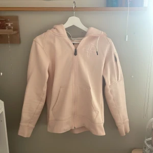Ljusrosa hoodie från Pelle P - Säljer en ljusrosa zip- hoodie från Pelle P med  broderad logga på bröstet. Tröjan har huva med snörning, fickor framtill och en patch på ärmen. Säljs då den bara hänger i garderoben. Jag köpte den för 350kr.