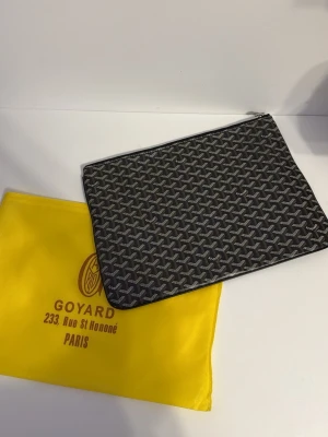 Svart Goyard kuvertväska med mönster - Snygg svart kuvertväska från Goyard med ikoniskt grafiskt mönster i grått och vitt. Väskan har dragkedja upptill och insida i starkt gul färg med en praktisk ficka. Perfekt för att bära dina essentials och ge din outfit en lyxig touch.