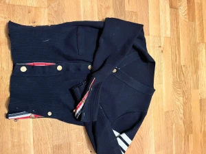 Mörkblå kofta med guldknappar - Snygg mörkblå kofta från Thom Browne med guldknappar och ribbad struktur. Har vita ränder på ena ärmen och coola detaljer med röd, vit och blå insida vid knappslån. Långärmad och med fickor framtill, perfekt för dig som gillar klassisk stil med twist.