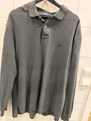 Grå långärmad polotopp från Polo Ralph Lauren - Säljer en stilren grå långärmad polotopp från Polo Ralph Lauren med klassisk krage och två knappar framtill. Toppens material är mjuk bomull och har en liten broderad logga på bröstet. Perfekt för dig som gillar enkel och clean stil.