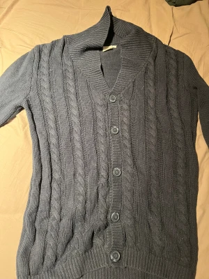 Mörkgrå kabelstickad kofta - Säljer en mörkgrå kabelstickad kofta från Lindex med bred krage och fem knappar framtill. Koftan har långärmad design och ribbade muddar vid ärmslut och nederkant. Perfekt för dig som gillar klassisk och mysig stil.