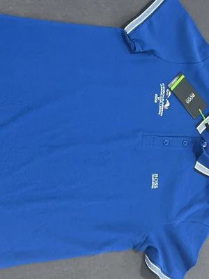 Blå piké från Hugo Boss Athleisure - Snygg blå pikétröja från Hugo Boss Athleisure med vit broderad logga och text på bröstet. Tröjan har korta ärmar med vit och blå randig mudd, klassisk krage och knappar framtill. Perfekt för dig som gillar sportig och stilren look.