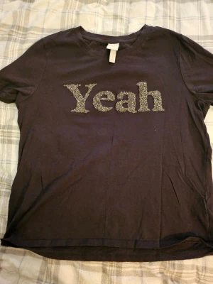 Svart t-shirt med glittrig text - Cool svart t-shirt från H&M med glittrigt 'Yeah'-tryck på bröstet. Klassisk rund hals och korta ärmar. Materialet är mjuk bomull som känns skönt mot huden. Perfekt för dig som gillar statement-plagg och vill sticka ut lite extra.