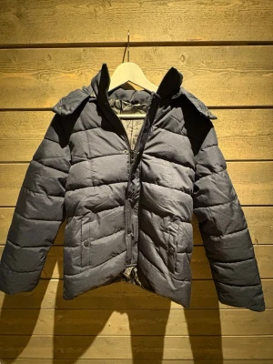 Mörkblå pufferjacka med huva - Säljer en mörkblå pufferjacka med huva och långa ärmar. Jackan har quiltad design, två fickor med knappstängning och dragkedja framtill. Perfekt för kalla dagar. Materialet är syntet och jackan är riktigt mjuk och skön. Jackan är aldrig använd.