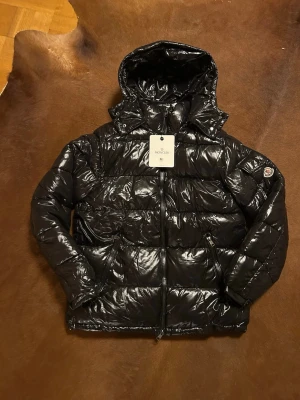 Svart Moncler pufferjacka med huva - Snygg svart pufferjacka från Moncler i glansigt material, med huva och ikonisk logga på ärmen. Jackan har dragkedja framtill och på fickorna, quiltad design och långärmad passform. Perfekt för dig som vill ha en stilren och varm jacka med premiumkänsla.
