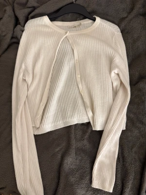 Ribbad vit kofta med knappar - Snygg ribbad vit kofta med rund hals och långa ärmar. Koftan har knappar framtill och är tillverkad i ett mjukt stickat material som känns skönt mot huden. Perfekt att slänga över en t-shirt eller linne för en chill look.