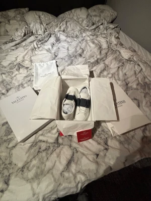 Valentino Opens blåa - Säljer ett par snygga Valentino Garavani Open sneakers i vitt skinn med bred blå läderdetalj över snörningen. Skorna har rund tå, platt sula med mönster och klassisk logga på tungan. Perfekt för dig som gillar lyxiga och stilrena sneakers.