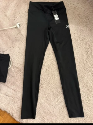 Svarta leggings från 157 - Svarta leggings från 157 med hög midja och stretchigt material. Snygg och enkel design med logga på låret, perfekta för träning eller chill. Tight passform som sitter skönt och följer kroppen.