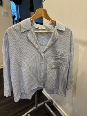 Randig blåvit skjorta från Zara - Säljer en luftig blåvit skjorta från Zara med tunna vertikala ränder, klassisk krage och bröstficka. Skjortan har knappar framtill och trekvartsärmar. Perfekt för en chill och stilren look.