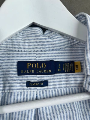 Randig skjorta från Polo Ralph Lauren - Klassisk blåvit randig skjorta från Polo Ralph Lauren med broderad logga på bröstet. Skjortan har lång ärm, knappar framtill och en tidlös button down-krage. Perfekt för dig som gillar en clean och stilren look. Fint skick, hittar inga fläckar, slitningar eller hål. 