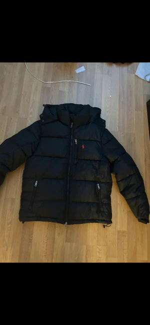 Svart pufferjacka från Polo Ralph Lauren - Svart pufferjacka från Polo Ralph Lauren med röd broderad logga på bröstet. Jackan har huva, två sidofickor med dragkedja och justerbar nederkant med snörning. Materialet är glansig polyester och jackan har en quiltad design som ger en schysst volym.