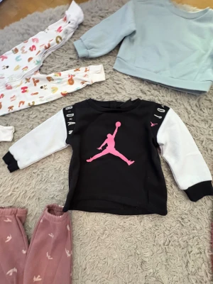 Jordan sweatshirt svart/vit rosa logo - Cool sweatshirt från Jordan med svart kropp, vita ärmar och stor rosa Jumpman-logga på bröstet. Texten 'JORDAN' i vitt på axlarna och små rosa loggor på ärmarna. Mjuk och bekväm tröja i bomull, perfekt för dig som gillar sportig streetstyle.