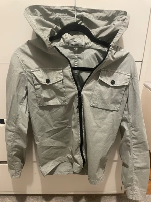 Stone Island Junior vindjacka grå - Snygg grå vindjacka från Stone Island Junior med huva, två stora bröstfickor med knappar och klassisk logotyp på ärmen. Jackan har dragkedja framtill. Perfekt för dig som gillar streetwear och clean stil.