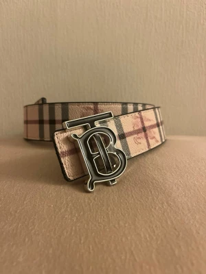 Rutigt bälte med TB-spänne - Snyggt bälte med klassiskt rutigt mönster i beige, rosa och grå toner. Bältet har ett metallspänne med Burberrys ikoniska TB-logga. Perfekt accessoar för att lyfta din outfit med en lyxig touch.