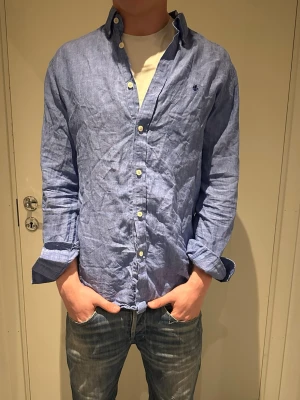 Blå linneskjorta från Morris  - Snygg blå skjorta i linne från Morris med klassisk button down-krage och vita knappar. Liten broderad logga på bröstet. Långärmad och avslappnad passform, perfekt till jeans eller shorts. Pris kan diskuteras vid snabb affär