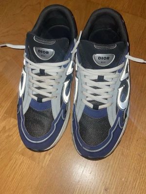 Dior B30 sneakers blå och silver - Säljer ett par Dior B30 sneakers i blått, svart och silver med reflekterande detaljer och vita snören. Skorna har en sportig vibe med mesh och syntetmaterial, samt en chunky sula för extra komfort. Perfekta för dig som vill sticka ut med lyxig streetstyle.