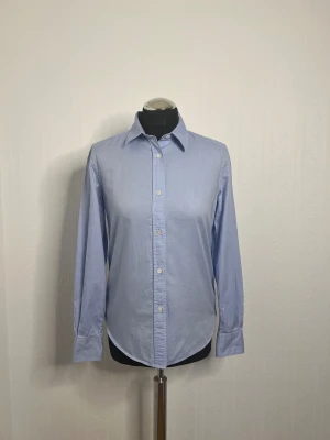 Ljusblå skjorta från ARKET - Snygg ljusblå skjorta från ARKET i klassisk modell med krage och knappar framtill. Skjortan har lång ärm och är tillverkad i mjuk bomull som känns skön mot huden. Perfekt för dig som gillar stilrena och enkla plagg med clean look. 100% bomull