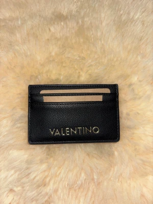 Valentino - Snyggt svart kortfodral från Valentino med guldfärgad logga framtill. Fodralet har flera fack för kort och är tillverkat i strukturerat skinn som ger en lyxig känsla. Perfekt storlek för att ha med sig i fickan eller väskan.