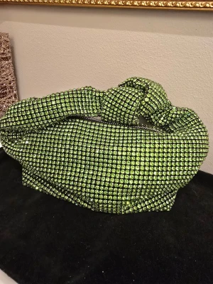 Grön glittrig handväska med knut - Unik handväska i glittrande grönt med massor av små stenar över hela ytan. Väskan har en cool knutdetalj på handtaget och en dragkedja upptill. Perfekt för dig som vill sticka ut och addera extra bling till din look.
