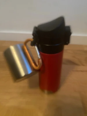 Termos och smidig kopp - Bilderna visar en röd termos med svart lock och en metallmugg fäst med en orange karbinhake. Ingen väst finns med på bilderna.