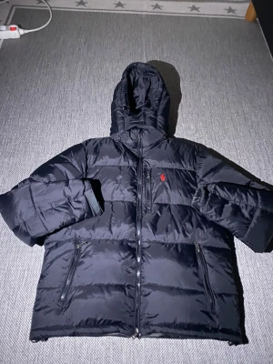 Svart pufferjacka från Polo Ralph Lauren - Svart pufferjacka från Polo Ralph Lauren med huva och dragkedja framtill. Jackan har två sidofickor med dragkedja och den klassiska röda Polo-loggan broderad på bröstet. Perfekt för kalla dagar och har en schysst, lite oversized look.