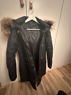 Svart lång Rockandblue dunjacka med fuskpäls - Säljer en svart lång dunjacka från Rockandblue med snygg quiltad design, huva med fuskpäls och dragkedja framtill. Jackan har även en liten ficka med dragkedja på ärmen och coolt Rockandblue-märke. Perfekt för kalla dagar!