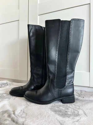 Svarta höga stövlar i skinn - Snygga svarta stövlar med rund tå och platt sula. Tillverkade i skinn med dekorativa detaljer längs skaftet och elastiska paneler på sidorna för extra komfort. Perfekta för dig som vill ha en stilren och klassisk look.