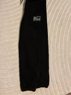 Svarta Stussy x Nike mjukisbyxor - Svarta mjukisbyxor från Stussy x Nike med broderad logga på benet. Byxorna har resår vid benslut och midja med snörning för skön passform. Perfekta för chill eller streetwear. Materialet är mjukt och bekvämt bomull.